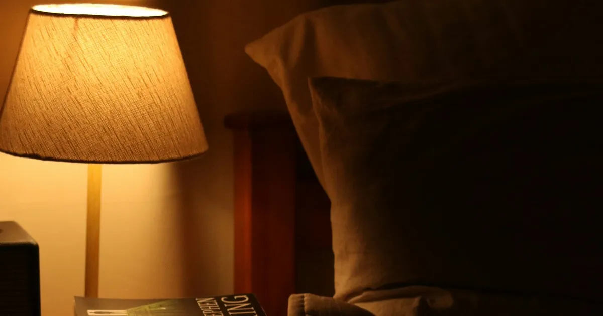 A lit bedside lamp glowing warmly in a dark bedroom, next to a pillow and headboard