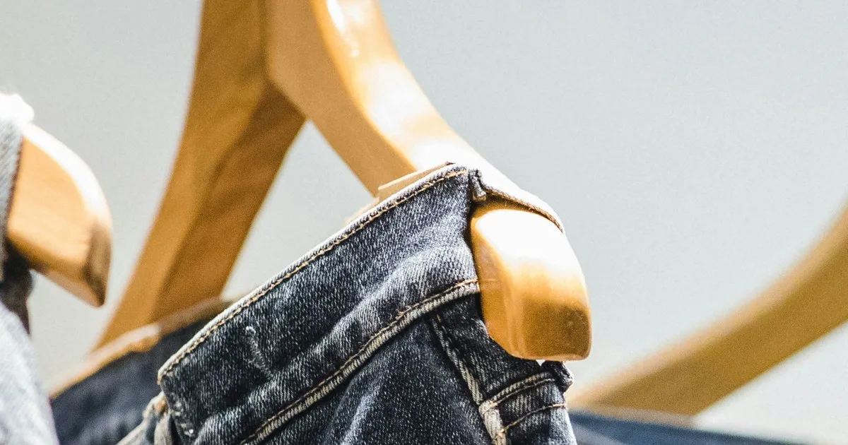 A denim waistband hanging on a wooden hanger against a plain wall